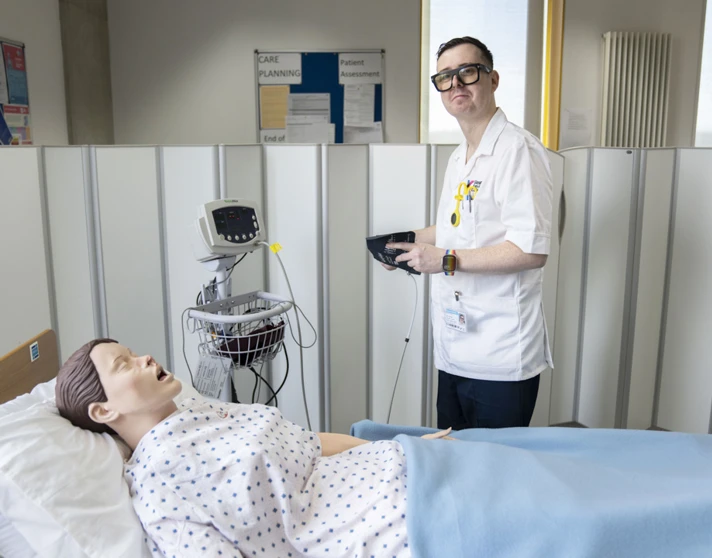 Student prepares to check vital signs on a training mannequin in a simulated hospital ward. Student prepares to check vital signs on a training mannequin in a simulated hospital ward.